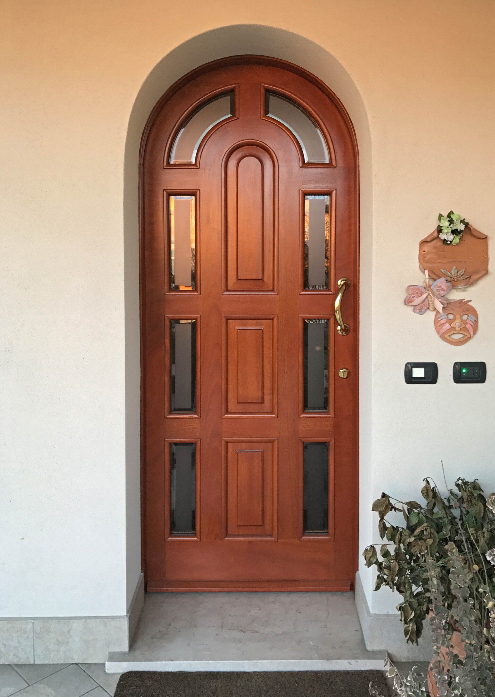 Una porta d'ingresso ad arco in legno con sei pannelli di vetro, incastonata in una parete di stucco chiaro con una piccola pianta sulla destra.