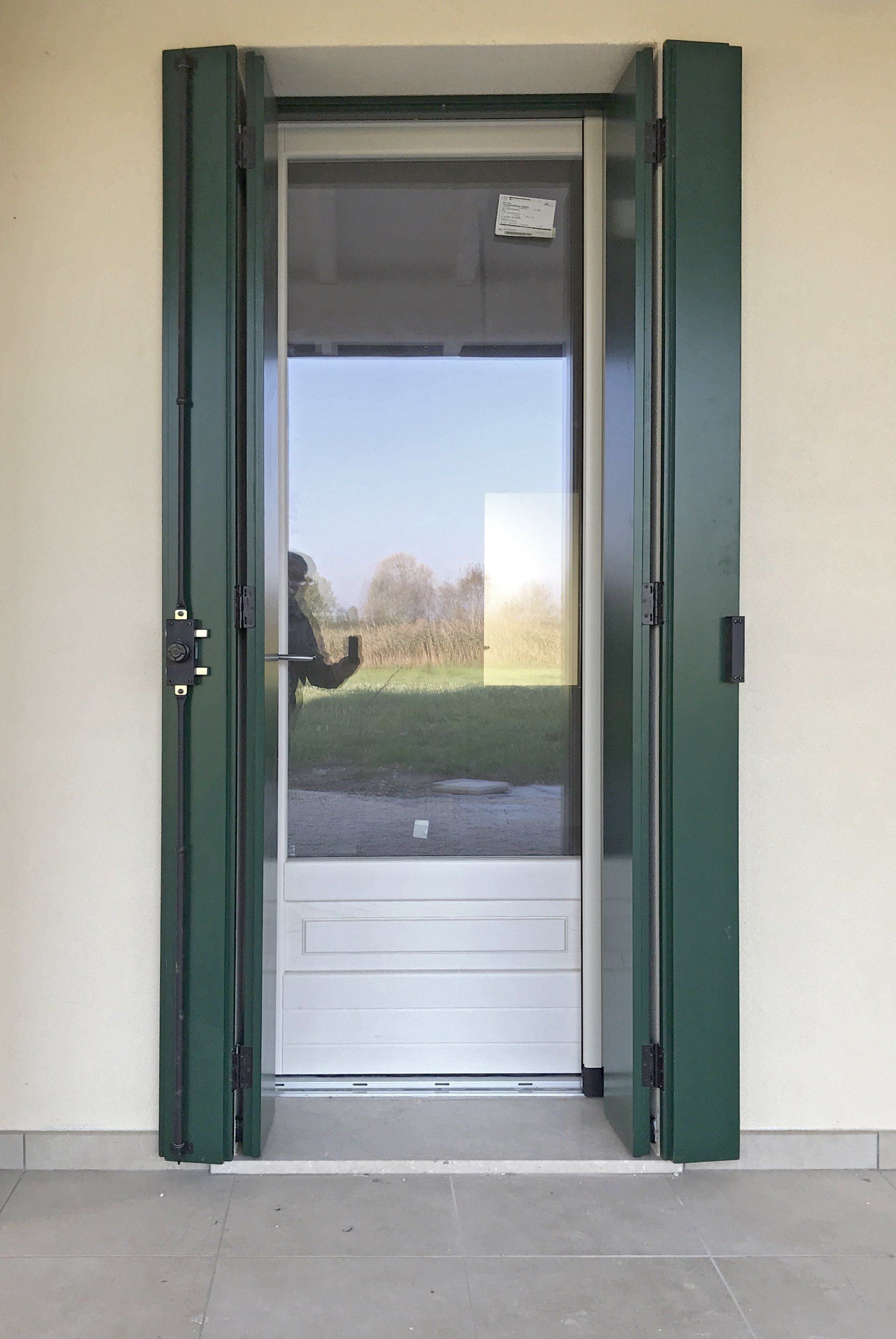 Una porta con pannelli di vetro bianco, incorniciata da persiane aperte di colore verde scuro, su una parete neutra.
