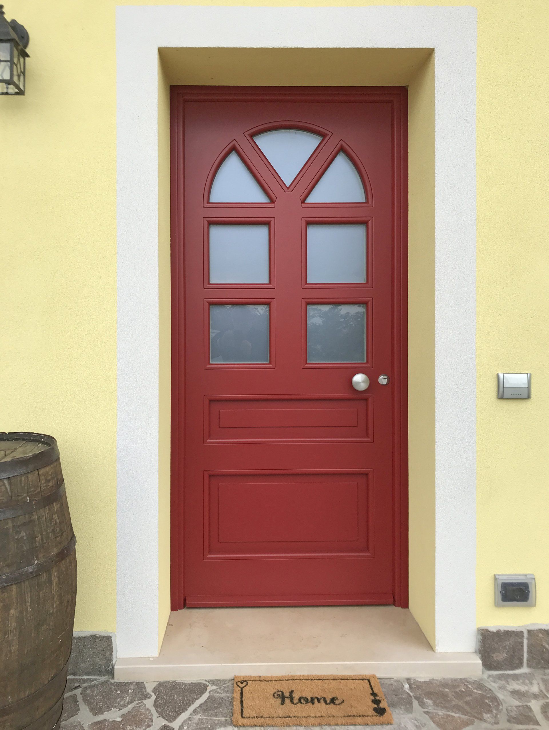 Una porta di legno rossa con pannelli di vetro incastonata in una parete gialla, con una botte di legno e uno zerbino con la scritta