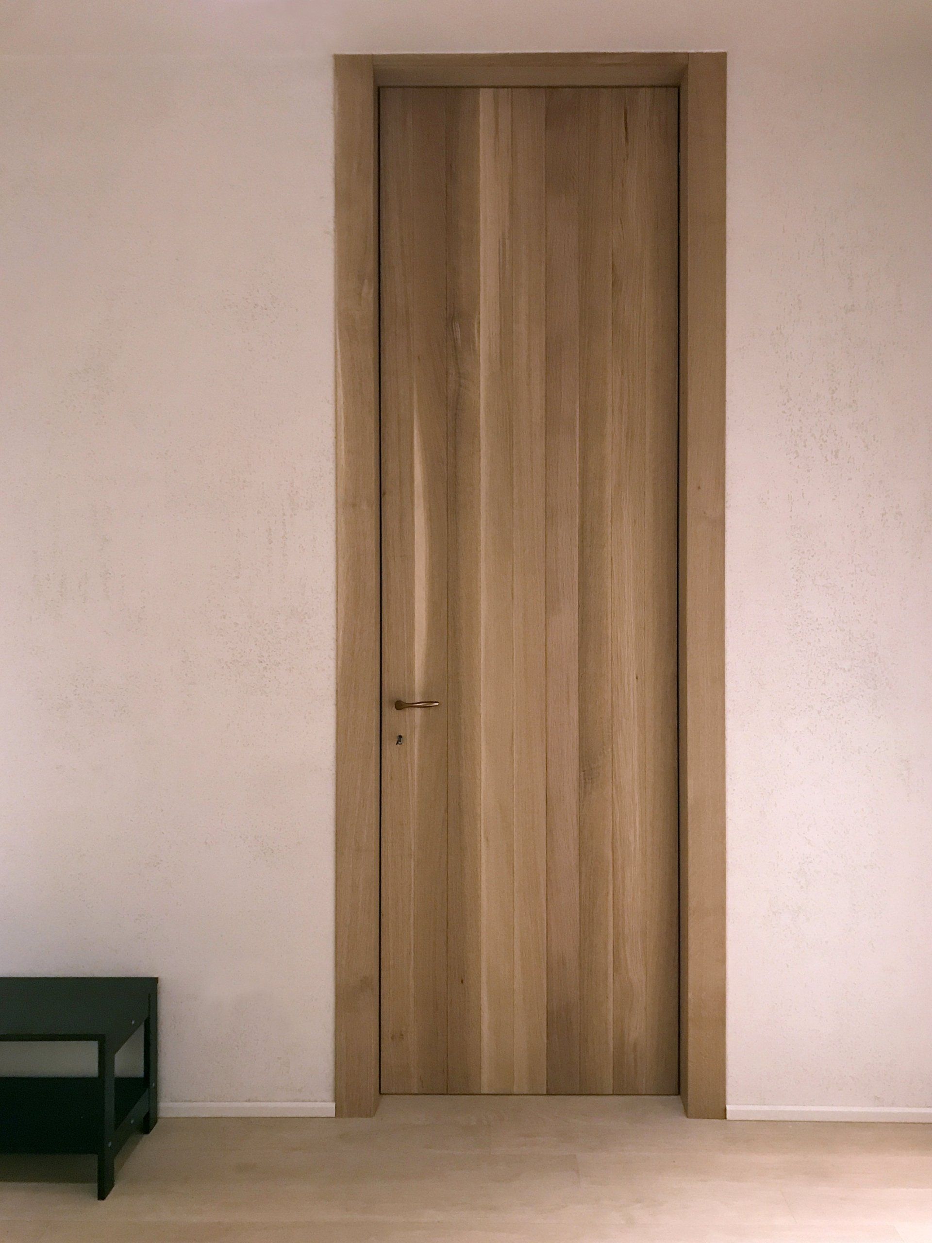 Una porta alta in legno con pannelli verticali incastonata in una parete bianca minimalista, accanto a un piccolo tavolino nero.