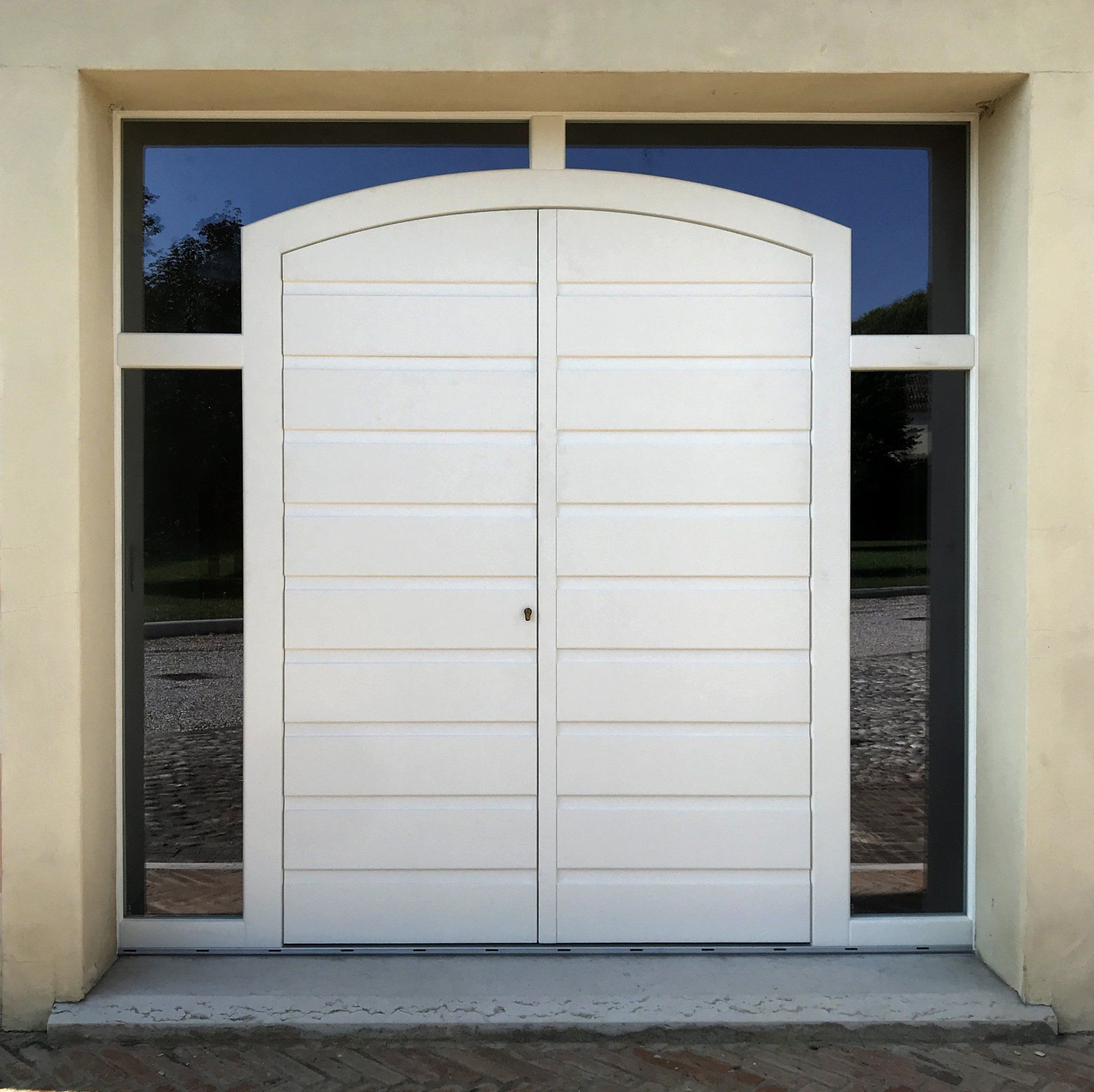 Una porta d'ingresso bianca a due ante con pannelli orizzontali, parte superiore ad arco e finestre laterali incastonate in una parete liscia.