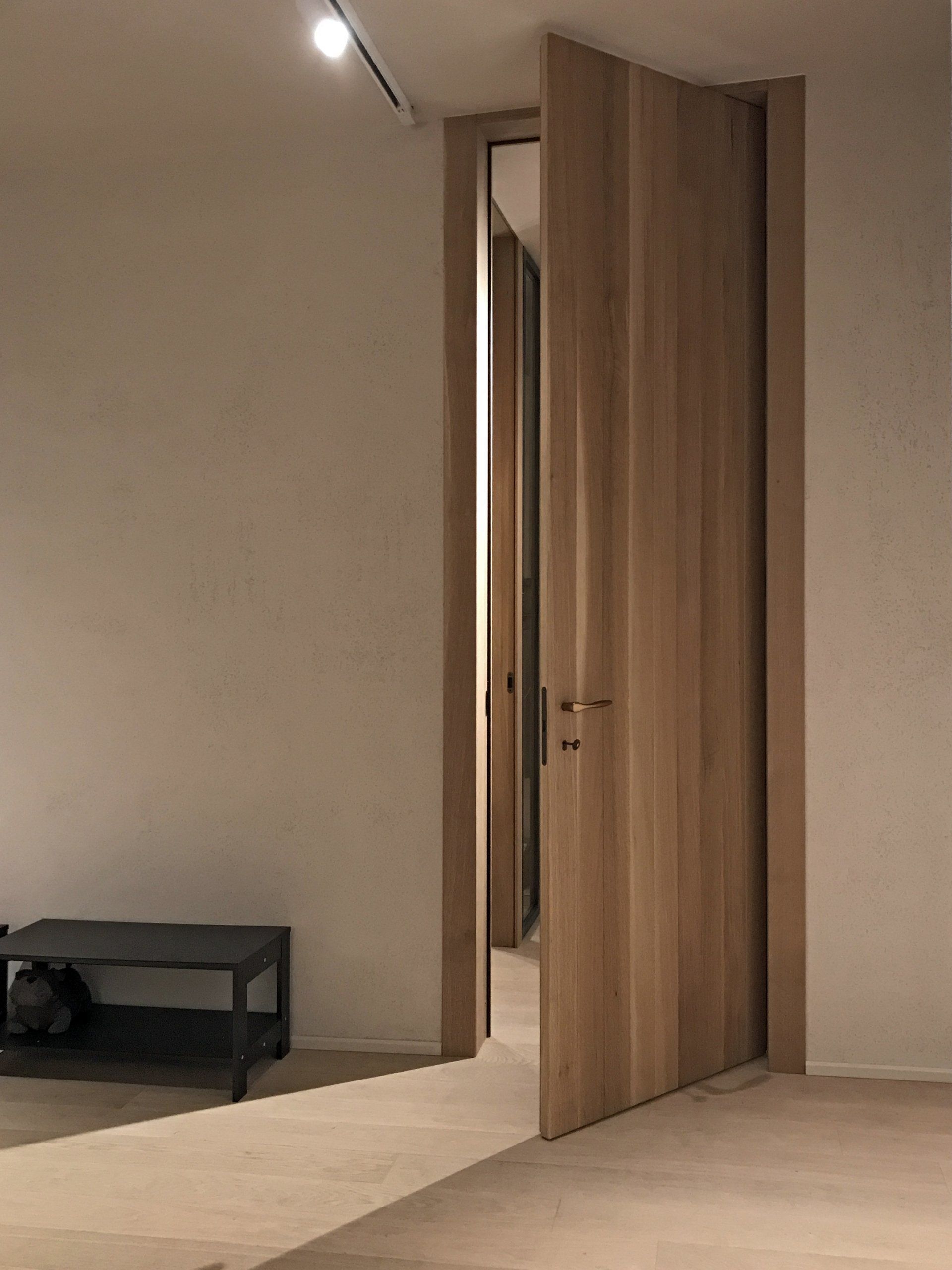 Un'alta porta di legno chiaro è leggermente socchiusa e si apre su un corridoio scarsamente illuminato, con una piccola panca scura in primo piano.