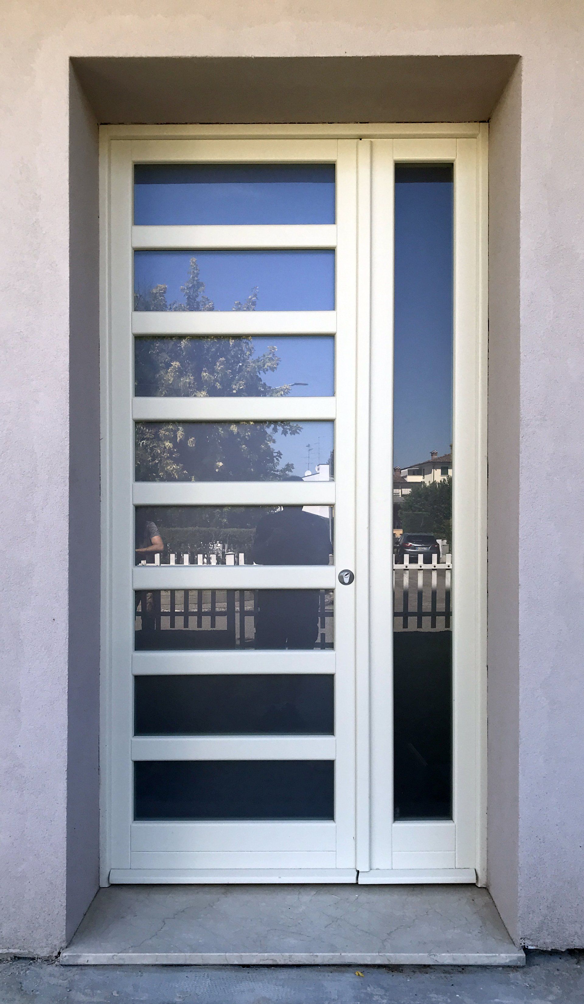 Una moderna porta d'ingresso color bianco sporco con sette pannelli di vetro orizzontali accanto a un unico pannello laterale verticale in vetro a tutta altezza.