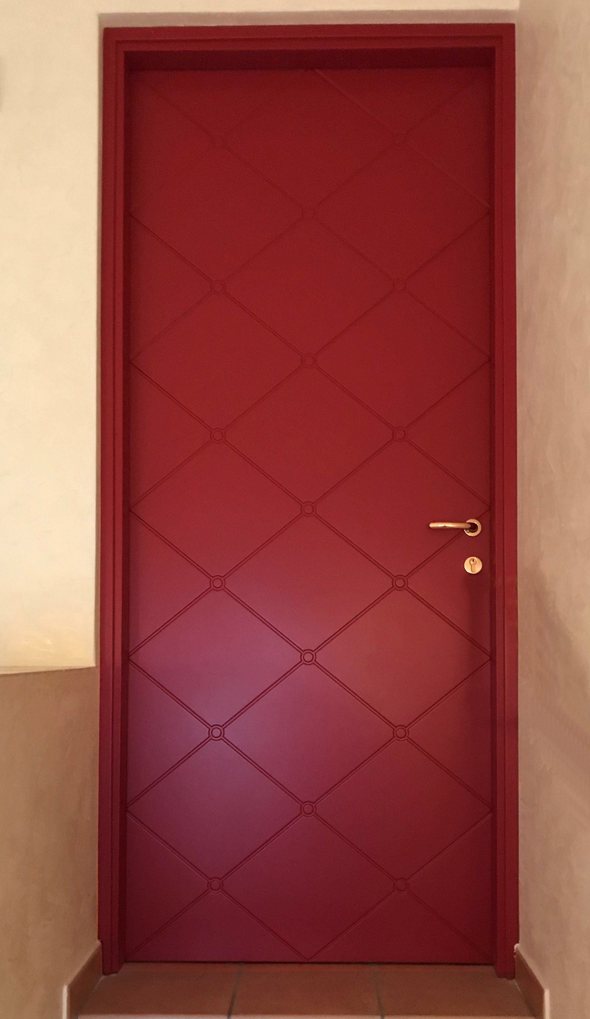 Una porta alta e rettangolare, dipinta di rosso scuro con un motivo a rombi in rilievo e una maniglia dorata, situata tra pareti beige.
