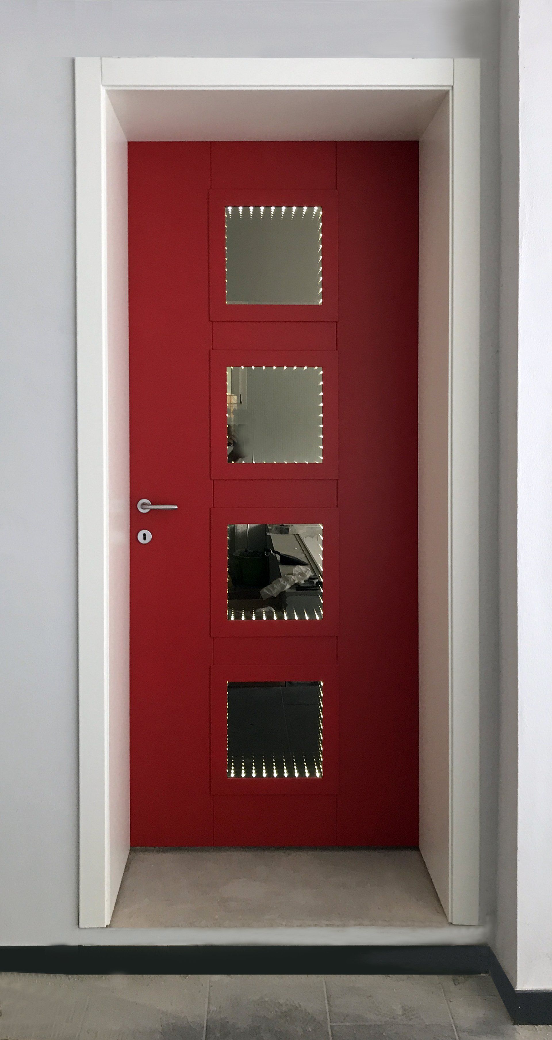Una porta rosso acceso con quattro pannelli di vetro quadrati equidistanti, incastonati in una cornice bianca su una parete grigia.