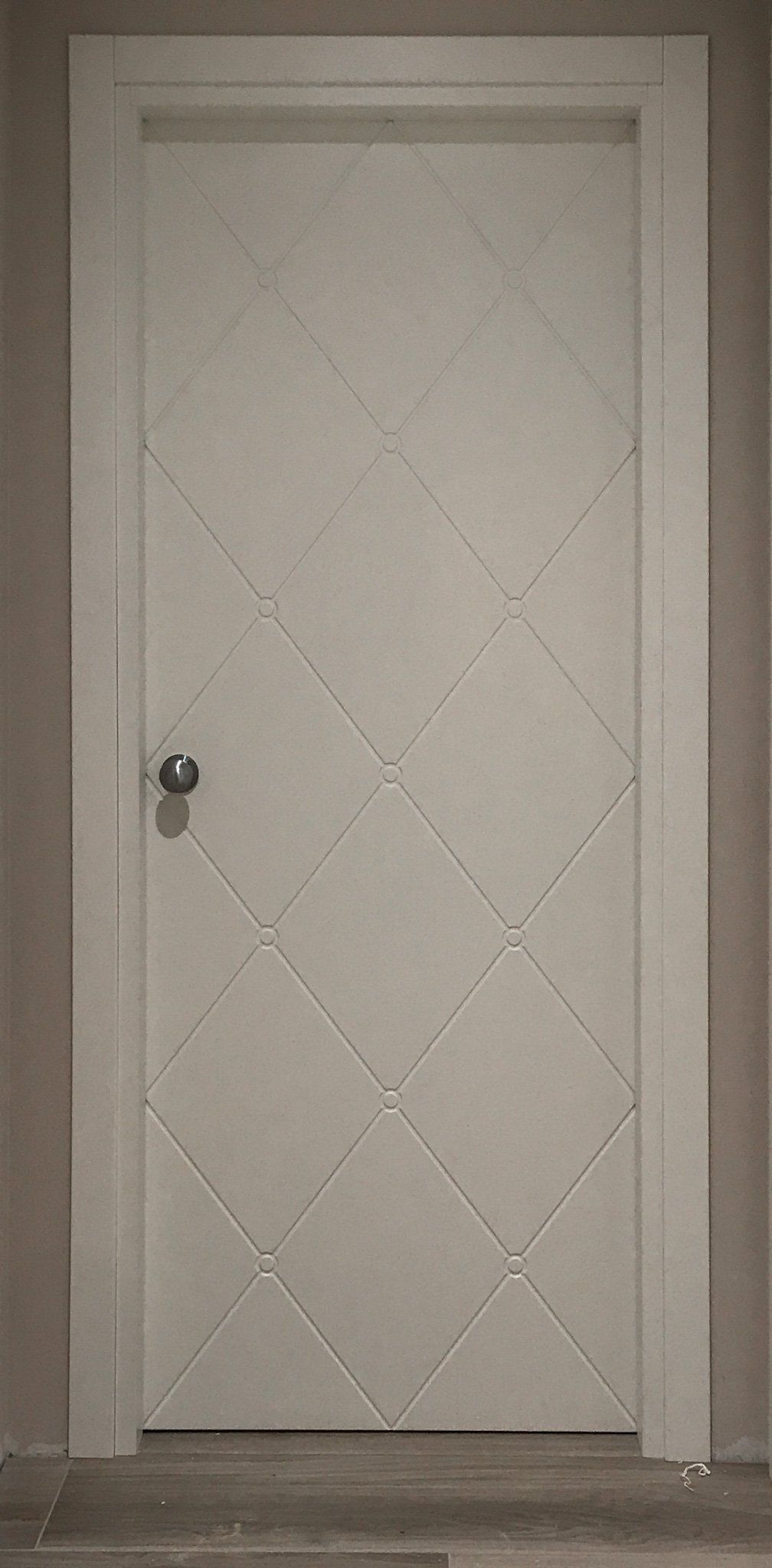 Una porta interna di colore chiaro con una trama trapuntata a rombi e dettagli a bottoni, dotata di maniglia rotonda.