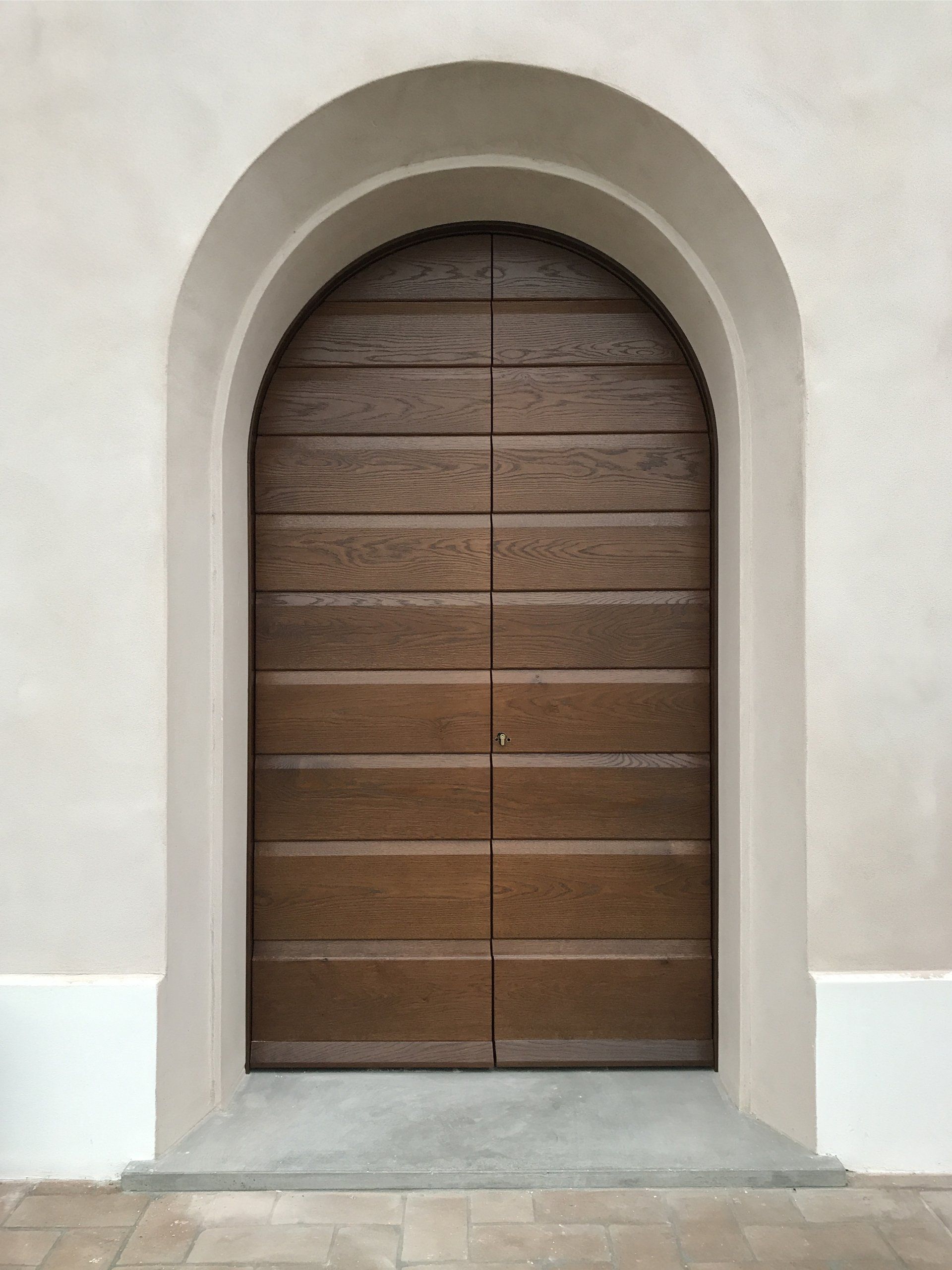 Una porta alta e arcuata in legno marrone con assi orizzontali, incastonata in un semplice muro di pietra chiara.