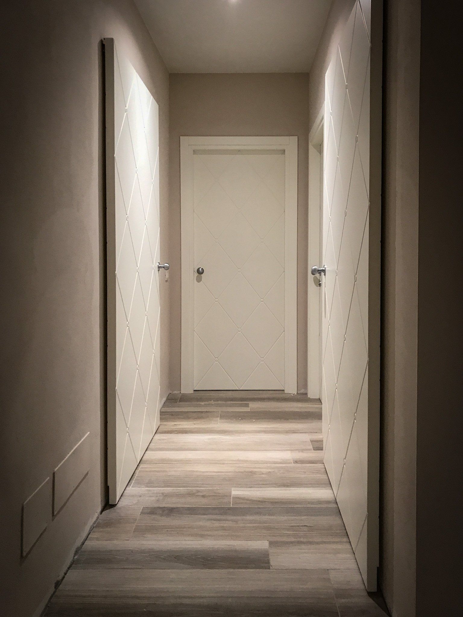Uno stretto corridoio interno con pavimento in legno chiaro, pareti beige e tre porte bianche con borchie metalliche decorative.
