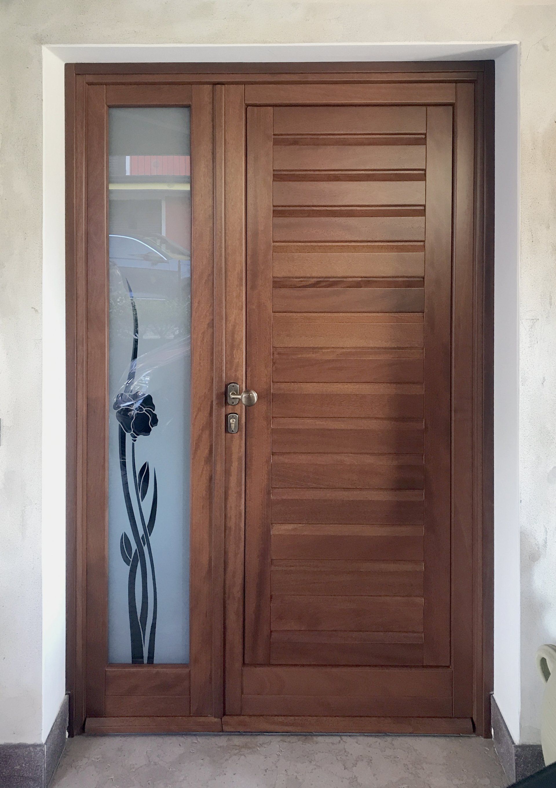 Una porta d'ingresso in legno con doghe orizzontali accanto a un pannello laterale con una finestra in vetro smerigliato decorata con motivi floreali.