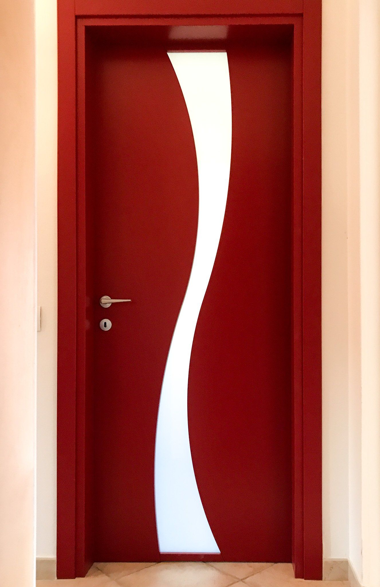 Una porta rossa con un pannello in vetro satinato verticale a forma di S, maniglia argentata e cornice semplice, vista da un corridoio.