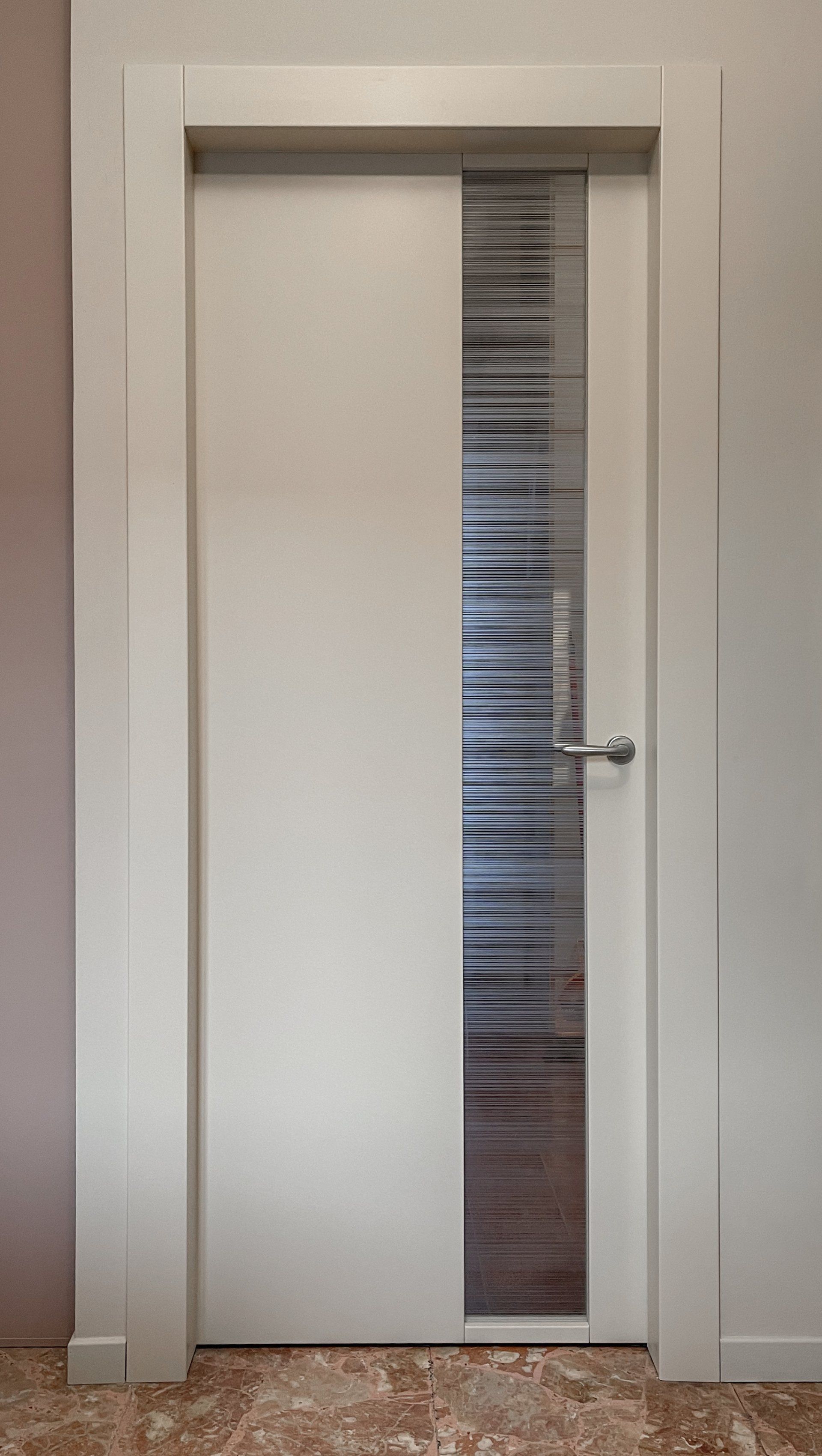 Una porta interna bianca con un pannello verticale in vetro satinato decorato con motivi a righe orizzontali e una maniglia argentata.