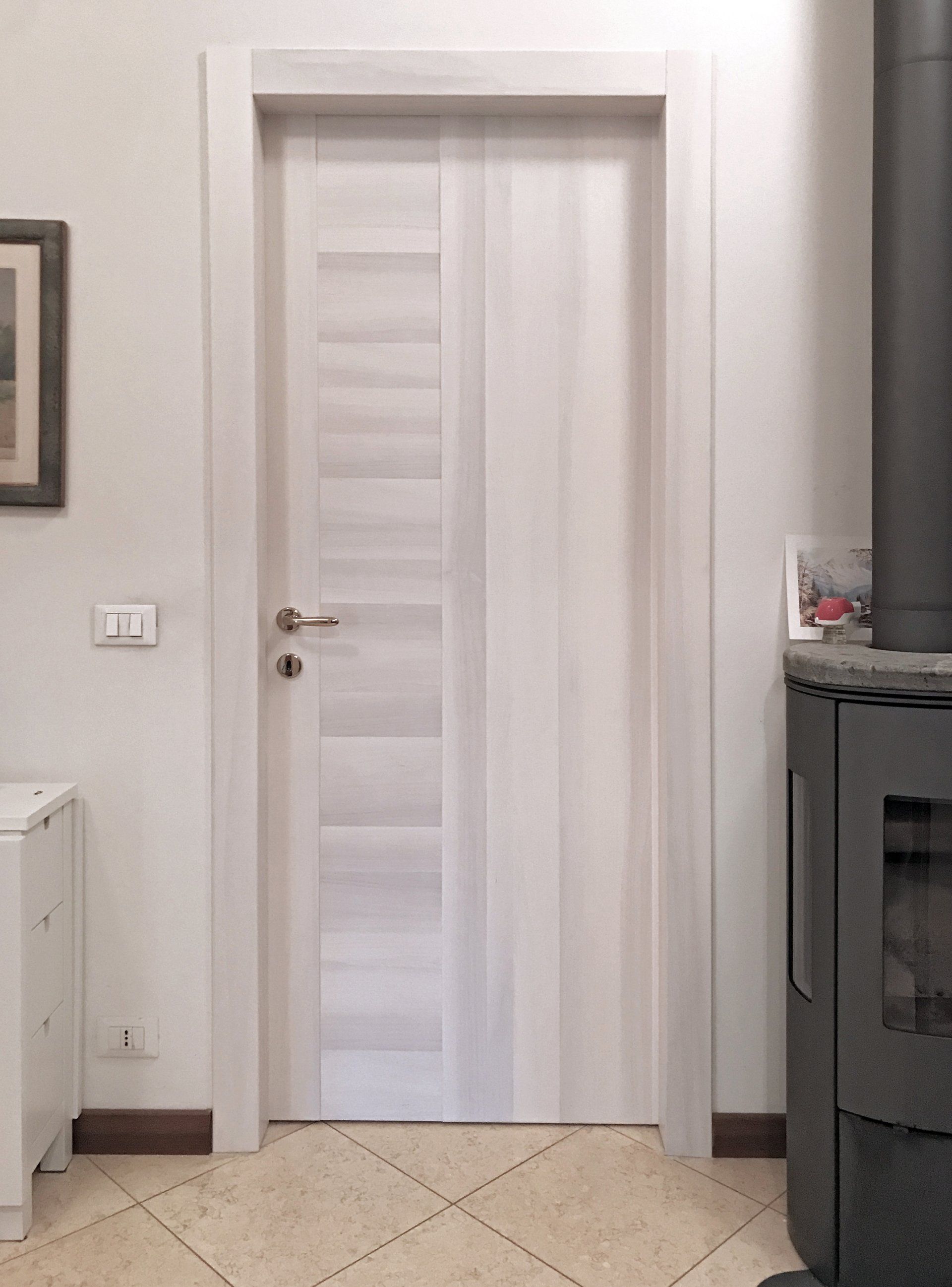 Una moderna porta interna in legno chiaro con maniglia argentata, incastonata in una parete bianca accanto a una stufa scura.