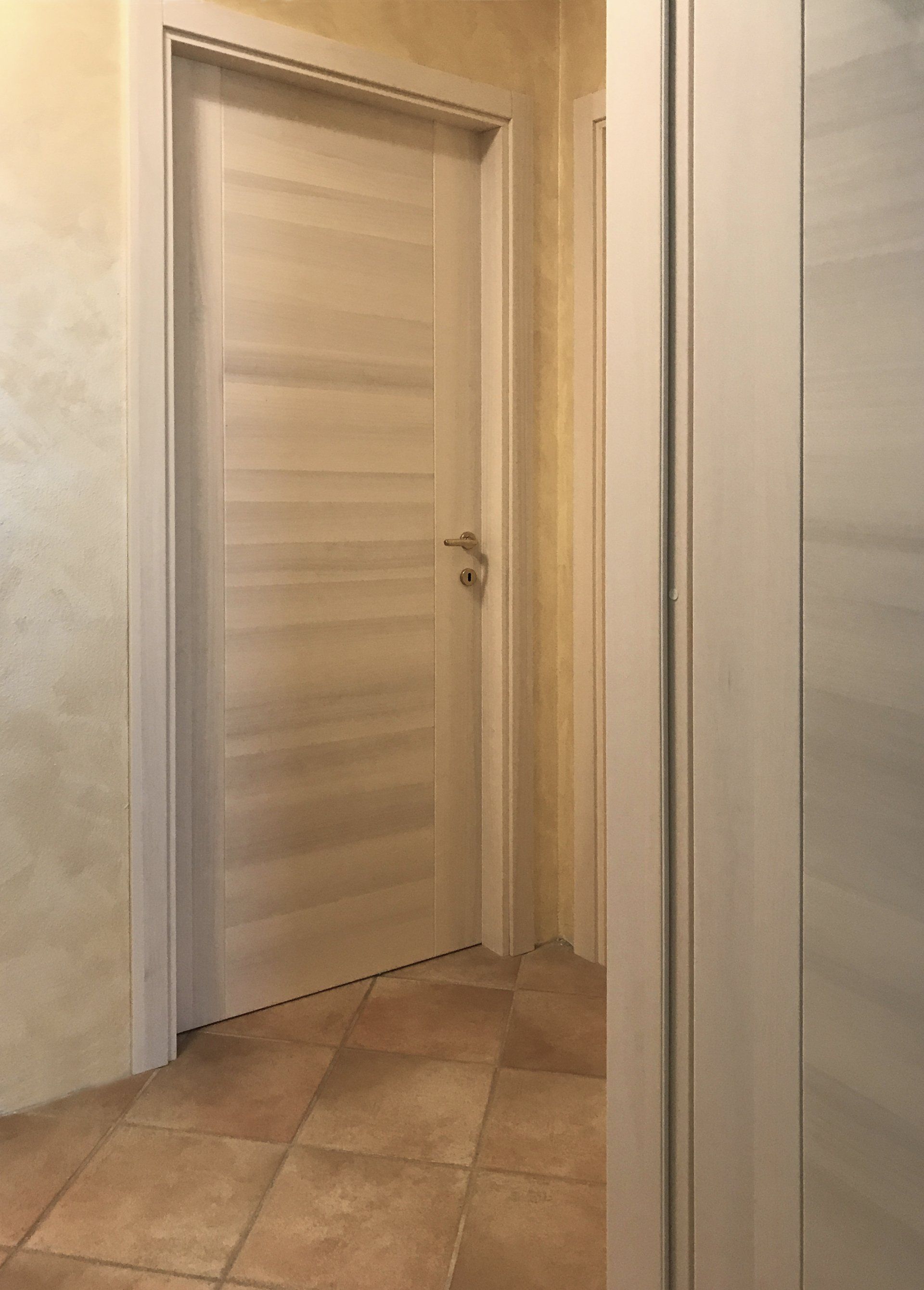 Una porta con venature del legno chiaro e maniglia argentata, inserita in un corridoio con pavimento piastrellato beige e pareti dai colori neutri.