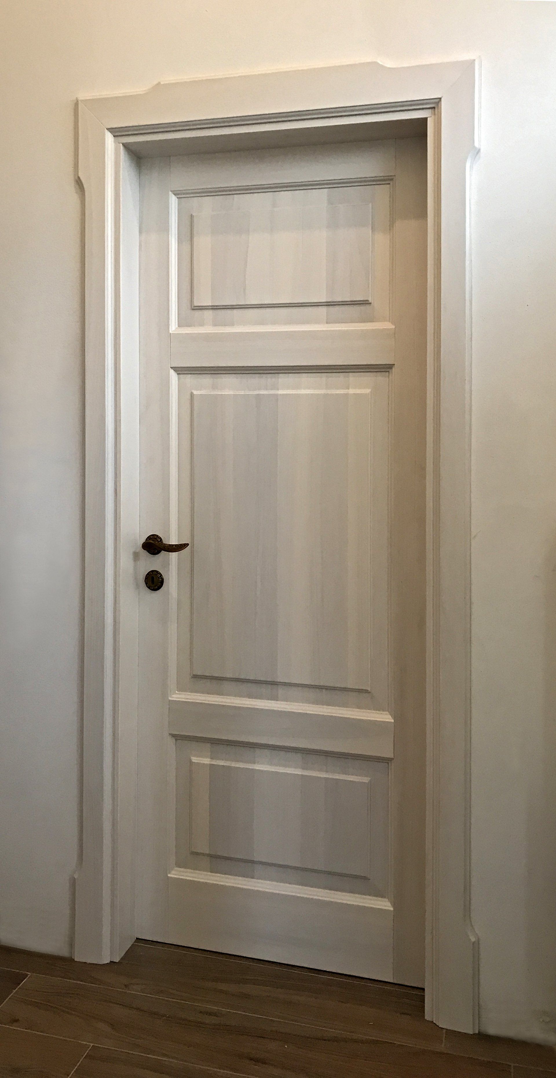 Una porta interna bianca a tre pannelli con ferramenta scura, incorniciata da una modanatura decorata, su una parete bianca.