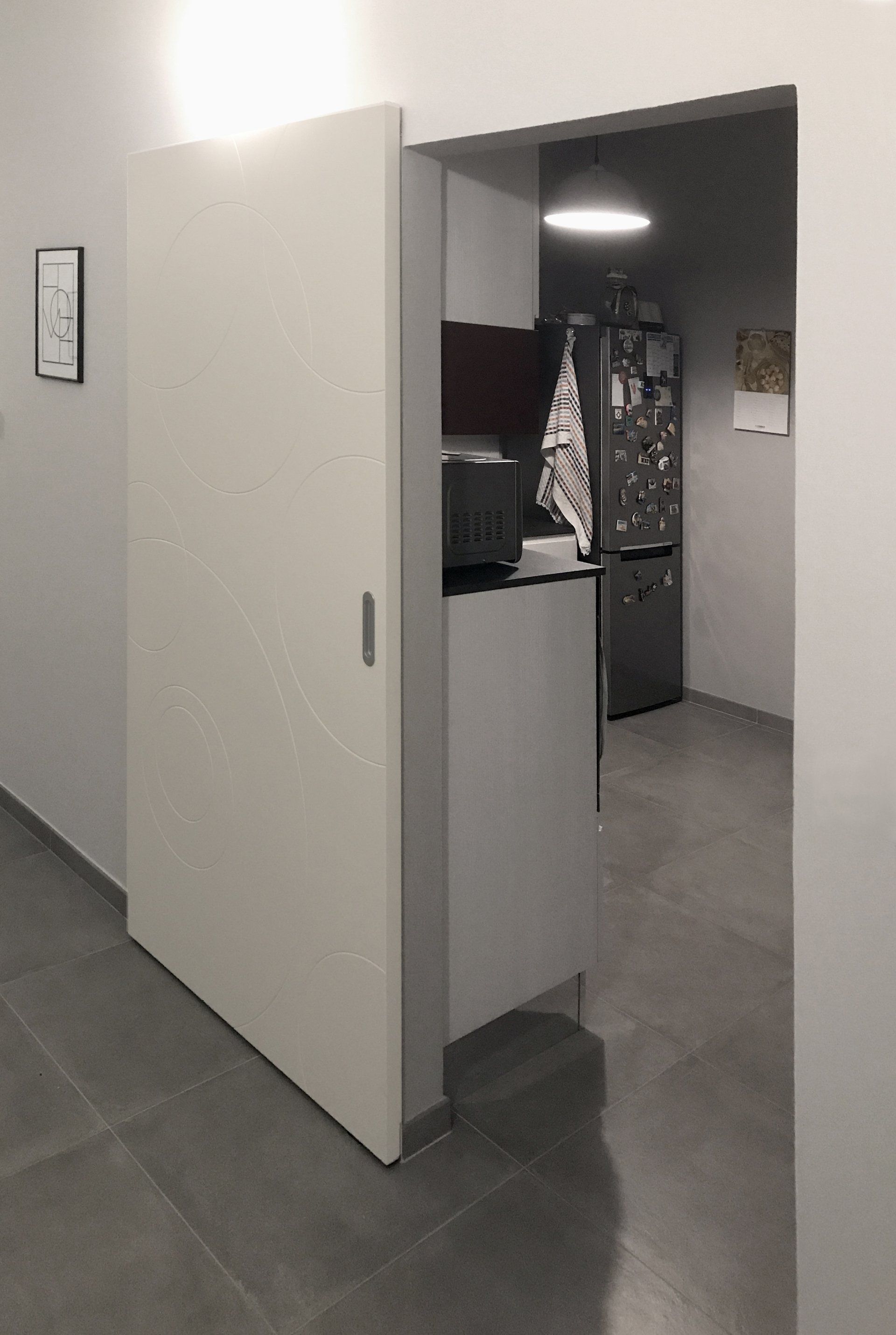 Una porta scorrevole bianca è parzialmente aperta, lasciando intravedere una cucina con pavimento grigio e frigorifero.