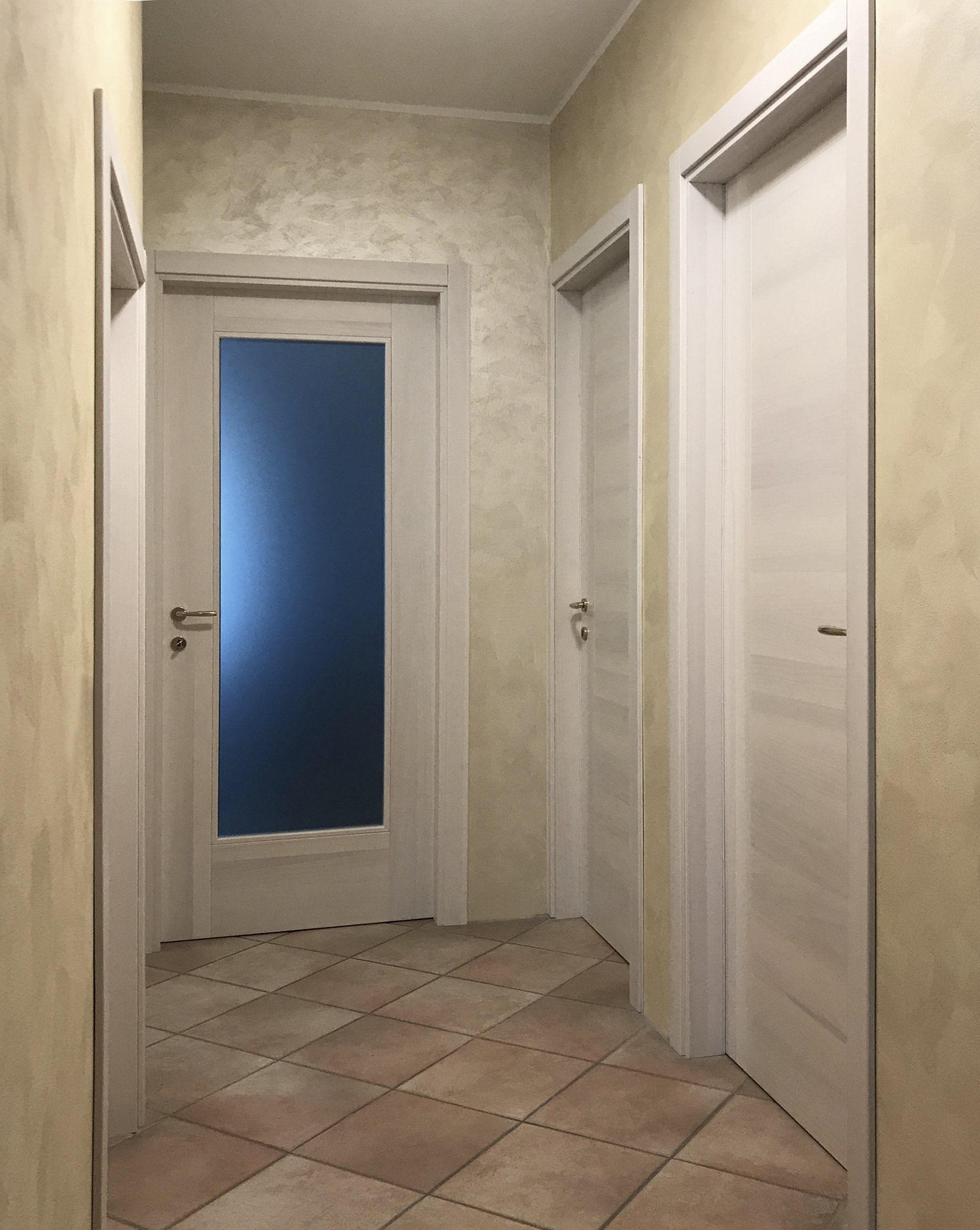 Un corridoio con pareti dalla texture chiara e pavimento in piastrelle color beige, caratterizzato da tre porte bianche chiuse, una delle quali con un pannello in vetro.