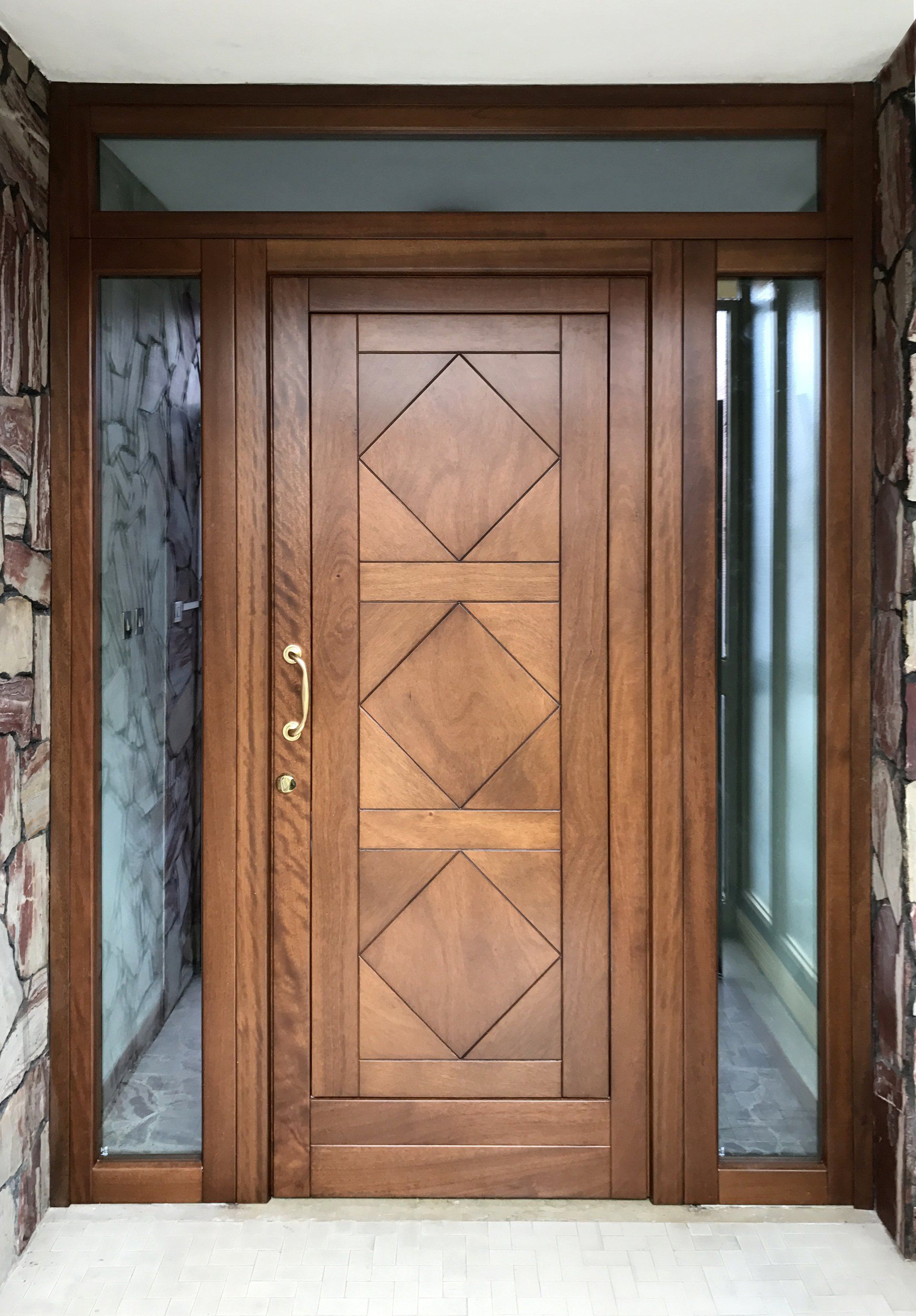 Una porta d'ingresso in legno con tre pannelli a forma di diamante, fiancheggiata da finestre laterali verticali in vetro e da una finestra a lunetta orizzontale.