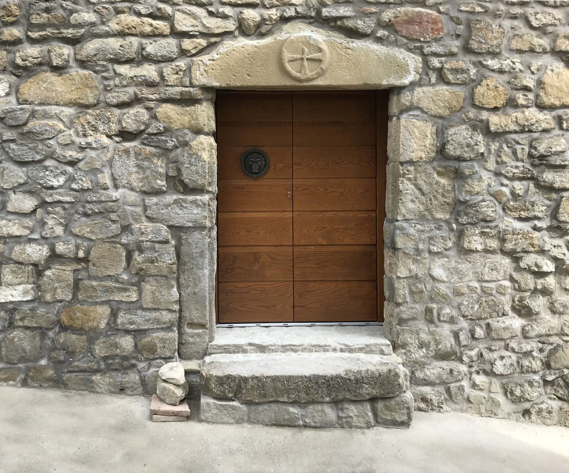 Una porta di legno incastonata in un muro di pietra, con un architrave in pietra sormontato da una croce scolpita e un piccolo gradino.