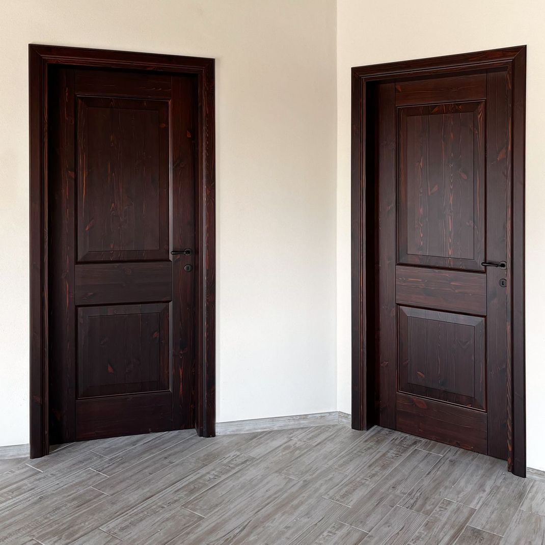 Due porte in legno scuro con modanature decorative su una parete bianca sopra un pavimento grigio effetto legno.