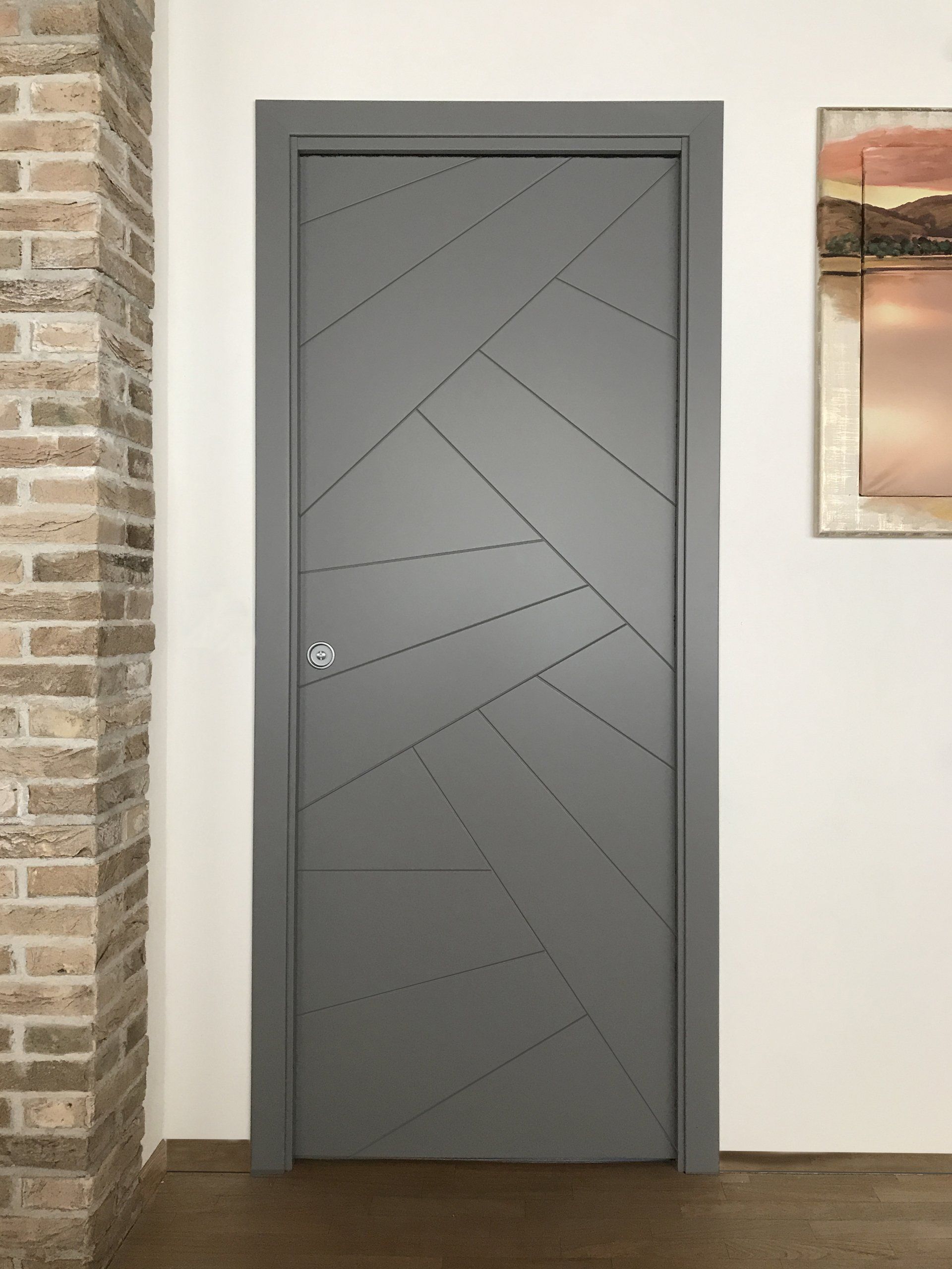 Una porta interna grigia con un moderno motivo geometrico a linee cucite, inserita in una stanza con una parete a vista in mattoni.