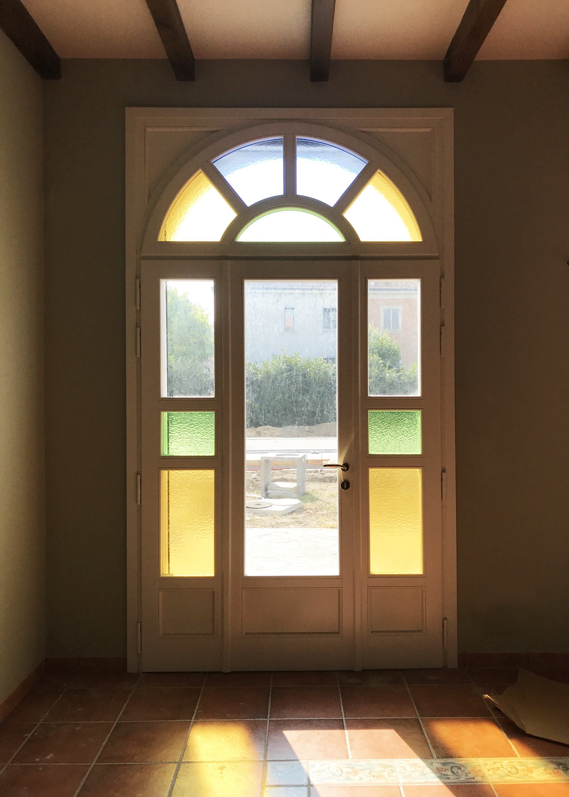 Vista interna di una porta in legno con sopraluce ad arco, pannelli in vetro e dettagli in vetro colorato di giallo.