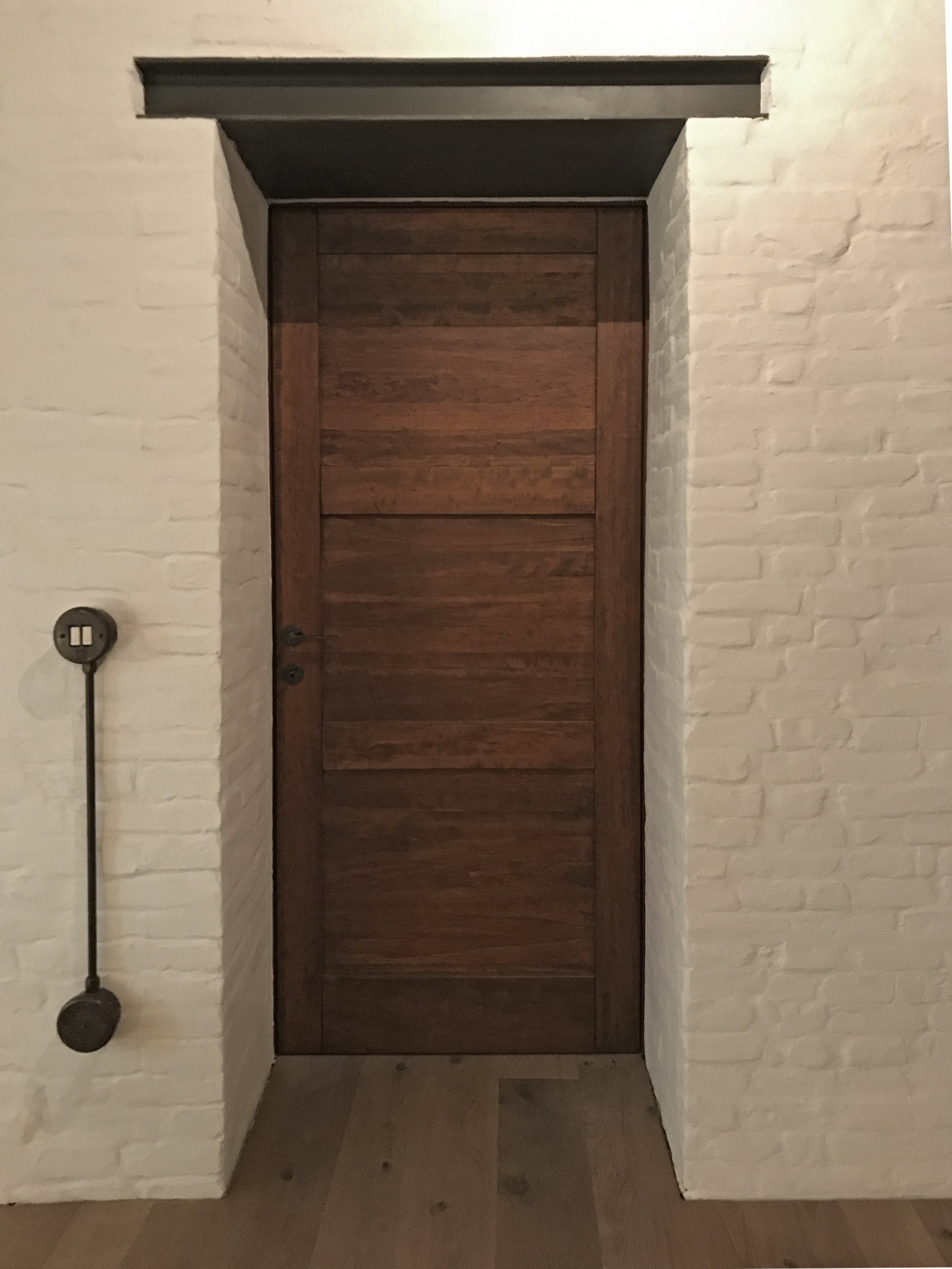 Una porta in legno scuro incastonata in un muro di mattoni dipinto di bianco, con un architrave in metallo sopra e un interruttore circolare a parete.