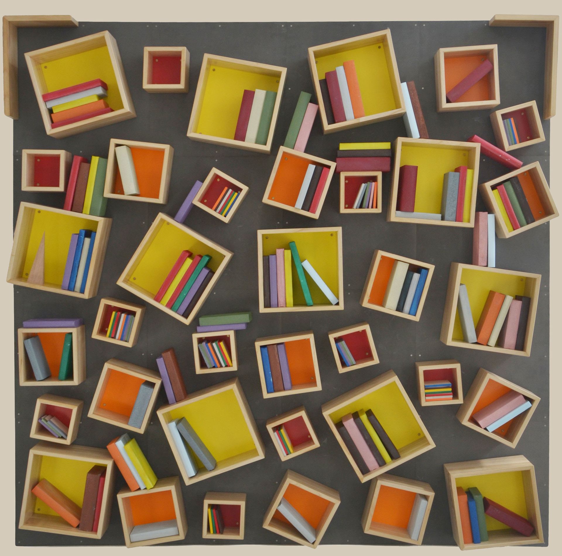 Una composizione a parete di mensole quadrate in legno, sparse e inclinate, che sorreggono colorati libricini in miniatura su uno sfondo grigio scuro.