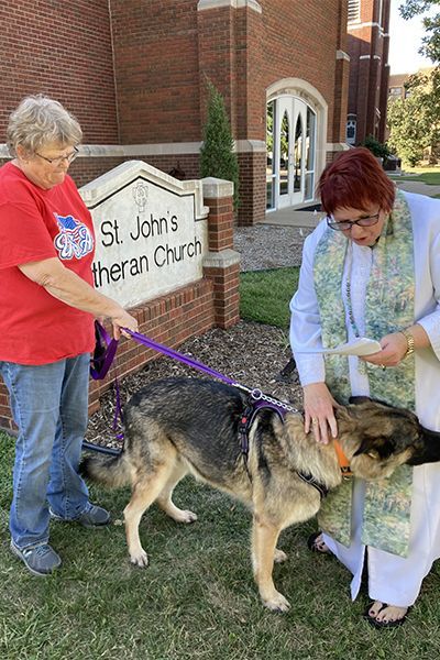 Two women are standing next to a dog on a leash being blessed in front of a church.