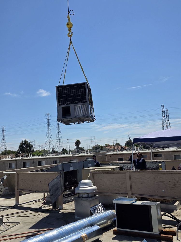 Crane lifting a large metal HVAC unit onto a rooftop under a clear blue sky