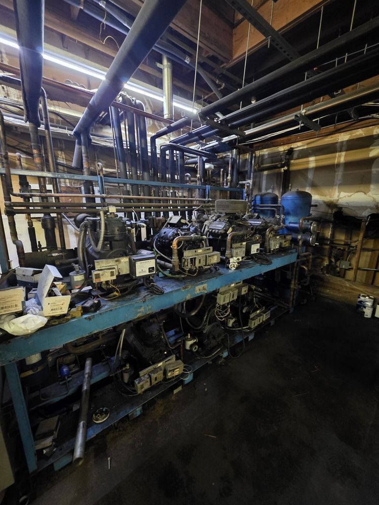 Industrial machinery and pipes in a dim factory room with blue shelving and equipment cluttered on workbenches