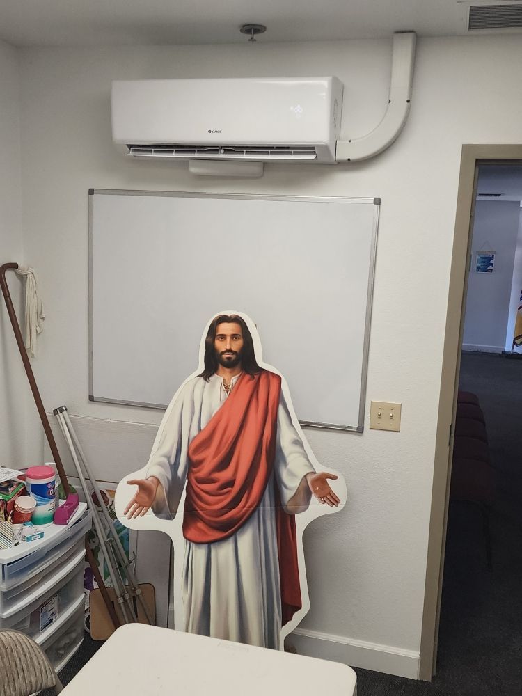 Cardboard cutout of Jesus standing in front of a whiteboard in a small room with an air conditioner above.