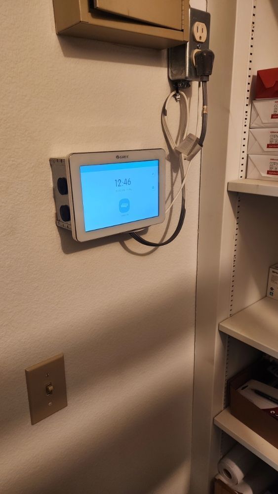 Blue wall-mounted touchscreen control panel with cables in a hallway or utility room.