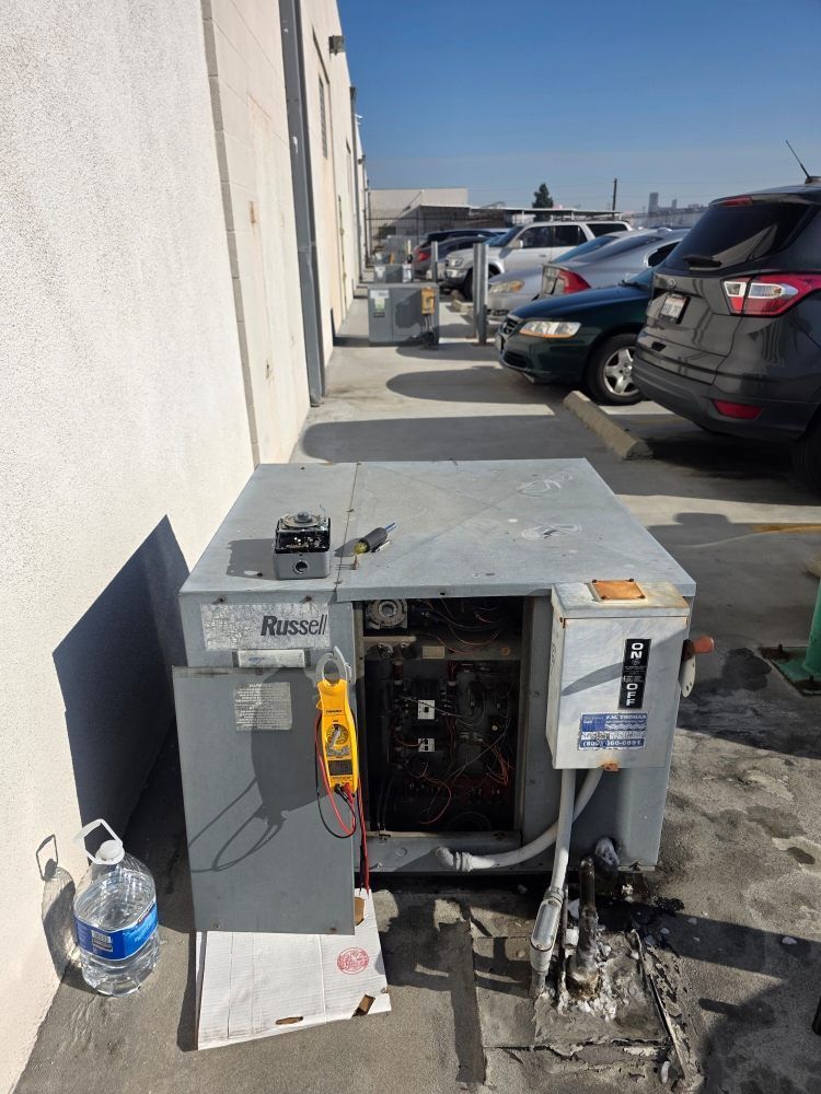 Damaged outdoor electrical unit beside a wall in a parking lot, with its panel open and debris around it