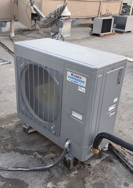 Gray outdoor air conditioning unit beside a building, with hoses attached on a rooftop.