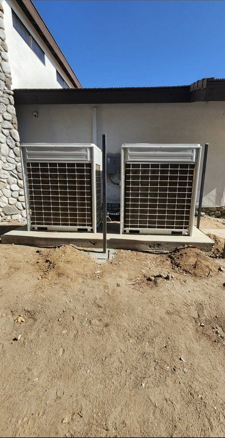 Two metal air conditioning units beside a building wall on bare dirt under a blue sky