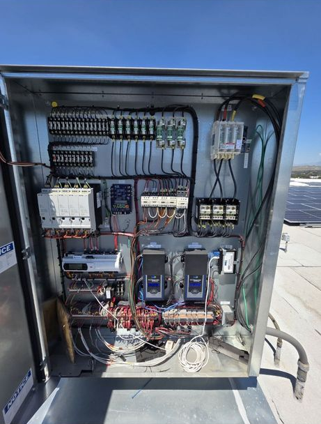 Open electrical control cabinet with wiring, breakers, relays, and blue drives on a rooftop.