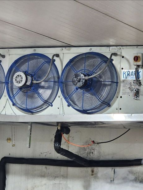 Two blue industrial fans mounted on a wall in a utility room, with pipes and wiring below
