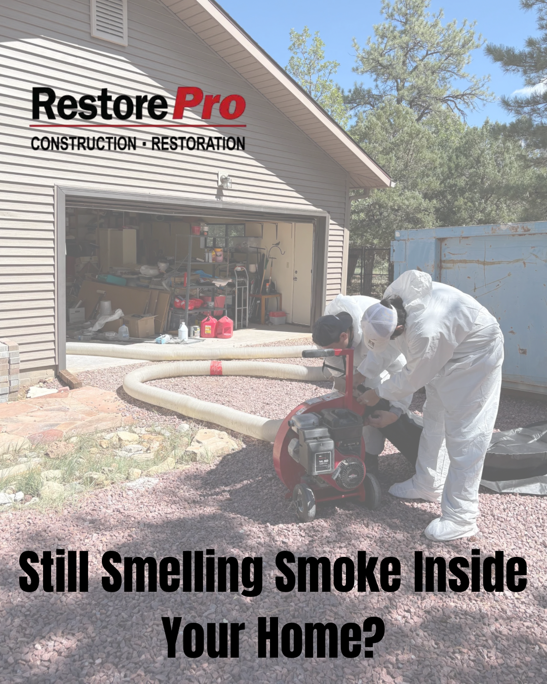 Worker in white protective suit using a saw outside a smoke-damaged house with garage open.