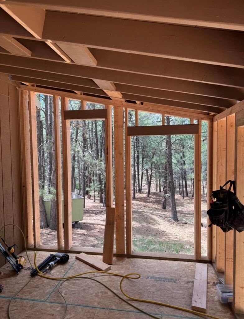 Unfinished wooden room interior with exposed beams, tools on floor, and forest visible through large window openings