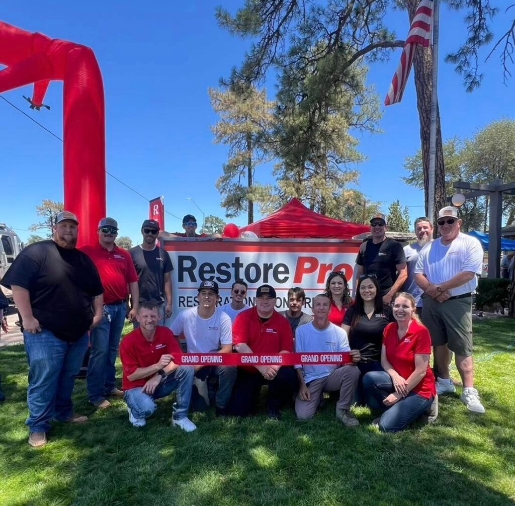 Group posing outdoors at a RestorePro event with red tents, banner, and team shirts under a blue sky Group posing outdoors at a RestorePro event with red tents, banner, and team shirts under a blue sky