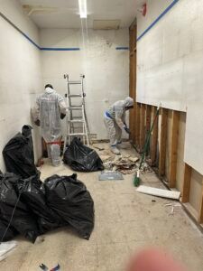 Workers in a narrow hallway clearing debris, with tools, ladders, and black trash bags spread around.