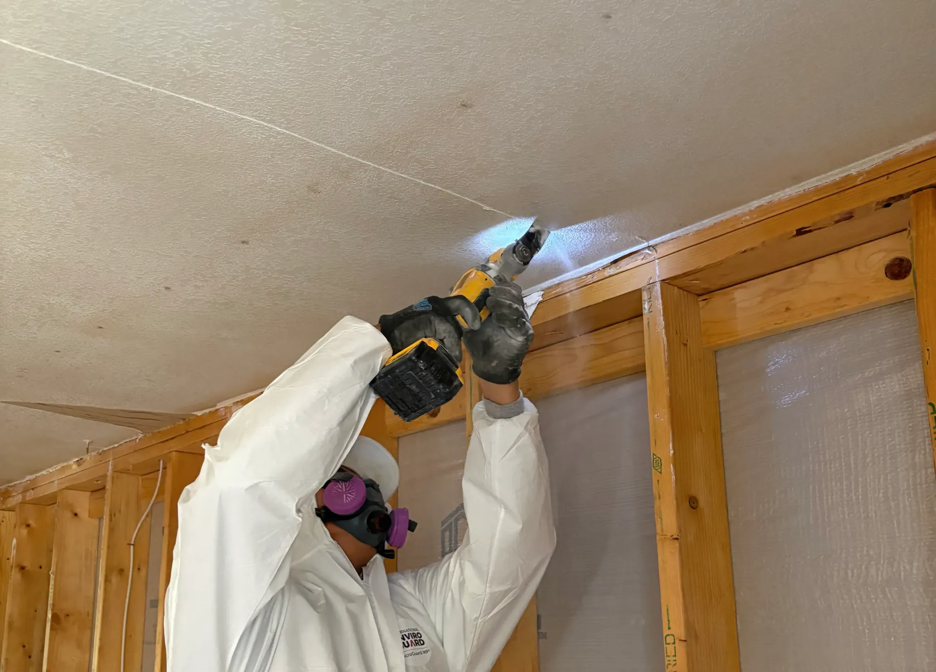 Person in white protective suit sprays foam sealant along a ceiling seam in an unfinished room