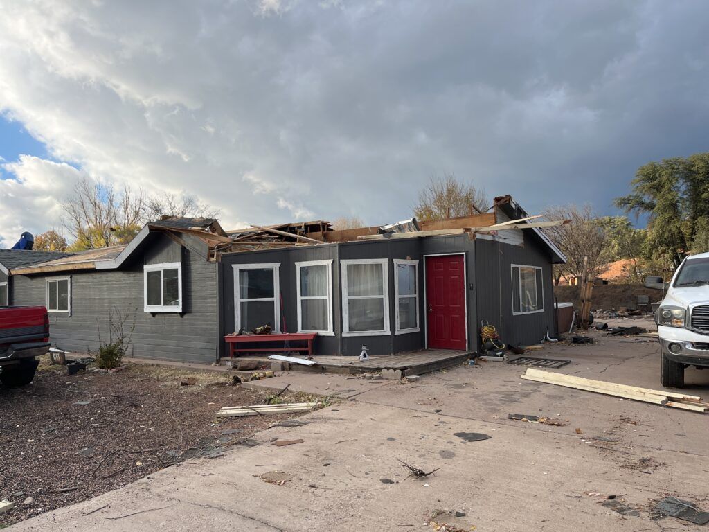 Damaged single-story house with a collapsed roof, red door, and debris in a driveway under cloudy skies