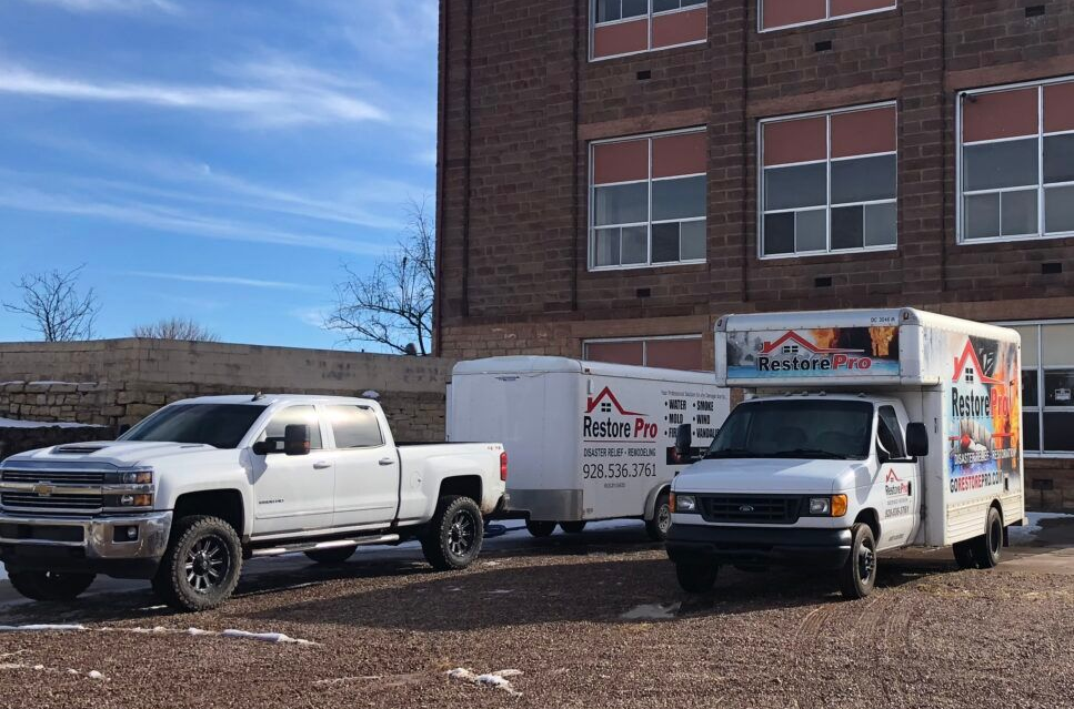 Two white utility trucks parked beside a brick building on a gravel lot, one with a moving trailer.