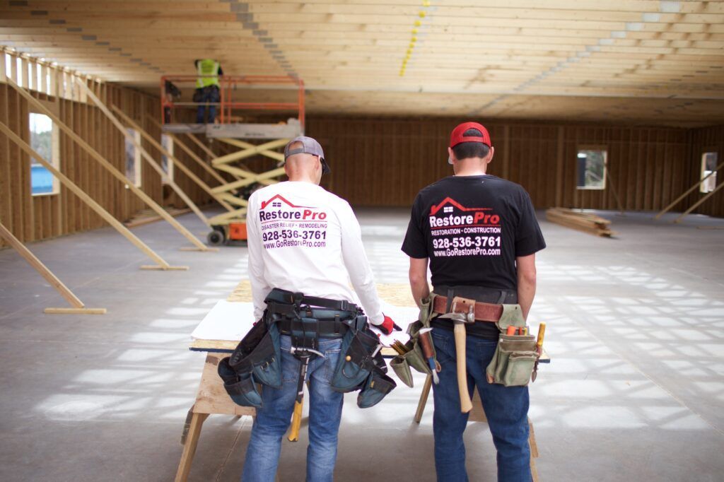 Two construction workers in a framed room, wearing tool belts and hard hats, walking away toward stairs.