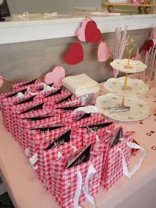 Pink checkered favor bags and a tiered dessert stand on a decorated party table with heart garlands.