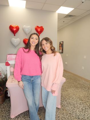 Two smiling people standing indoors beside heart balloons and a pink-covered chair