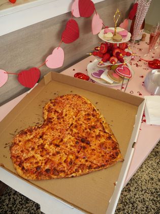 Heart-shaped pizza in a box beside pink and red heart decorations on a table