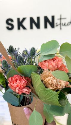 Bouquet of pink and tan flowers with green leaves in a brown paper wrap, in front of a SKNN studio sign