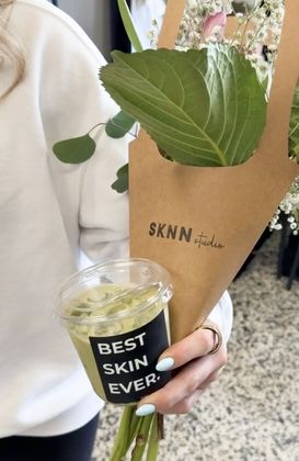 Person holding a cup labeled “BEST SKIN EVER” and a small wrapped bouquet with green leaves.